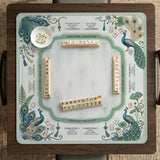 Teal square mahjong mat with peacock feathers, soft floral pattern, tassel details, and printed rule guide text in the center.