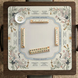 Beige square mahjong mat with a sage border, crane birds, bamboo, flowers, tile icons, and printed rules around a clean center play area.