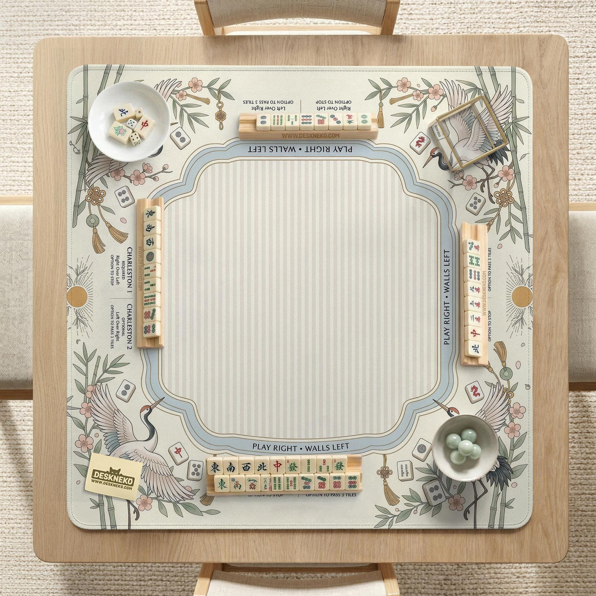 Beige square mahjong mat with a sage border, crane birds, bamboo, flowers, tile icons, and printed rules around a clean center play area.