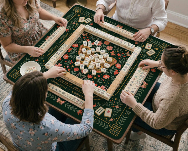 Dark botanical mahjong table mat with a floral border, large moth and butterfly design in the center, gold corner frame, and printed rules layout around the edge.