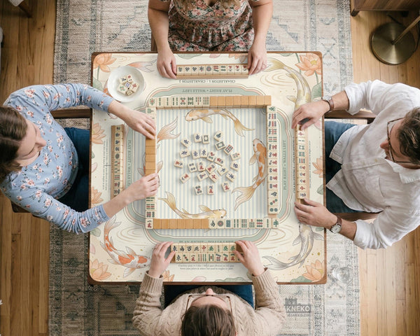 Sage and cream mahjong mat with a koi fish pond pattern, lotus blooms, water ripples, and printed instructions around the border. One version has a scalloped edge.