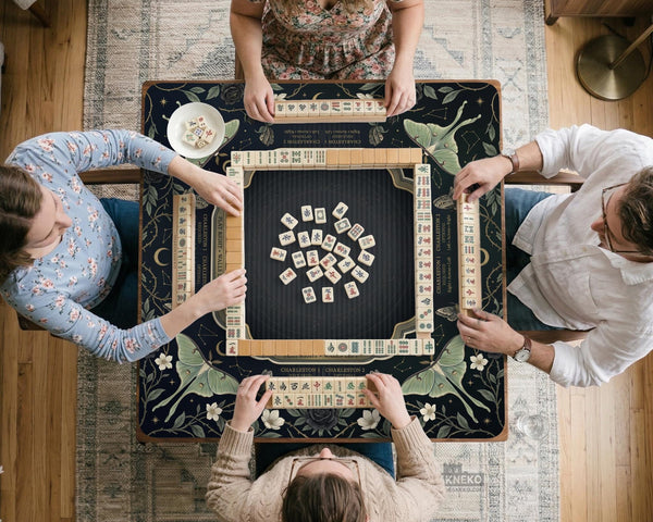 A dark floral mahjong mat with a scalloped edge, moth and flower artwork, and an American Mahjong rules layout on a dark background.