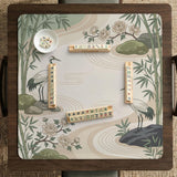 Square mahjong mat with a cream background, sage green cranes, bamboo leaves, peony branches, stones, and thin linework.