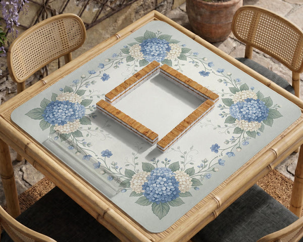 A square blue mahjong mat with hydrangea flowers, cream blossoms, sage leaves, and vines around a plain center play area. The edge is scalloped.