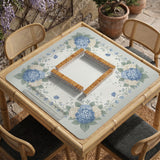A square blue mahjong mat with hydrangea flowers, cream blossoms, sage leaves, and vines around a plain center play area. The edge is scalloped.