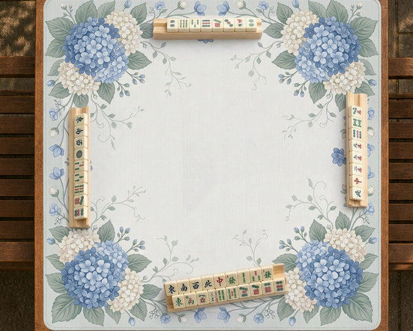 A square blue mahjong mat with hydrangea flowers, cream blossoms, sage leaves, and vines around a plain center play area. The edge is scalloped.