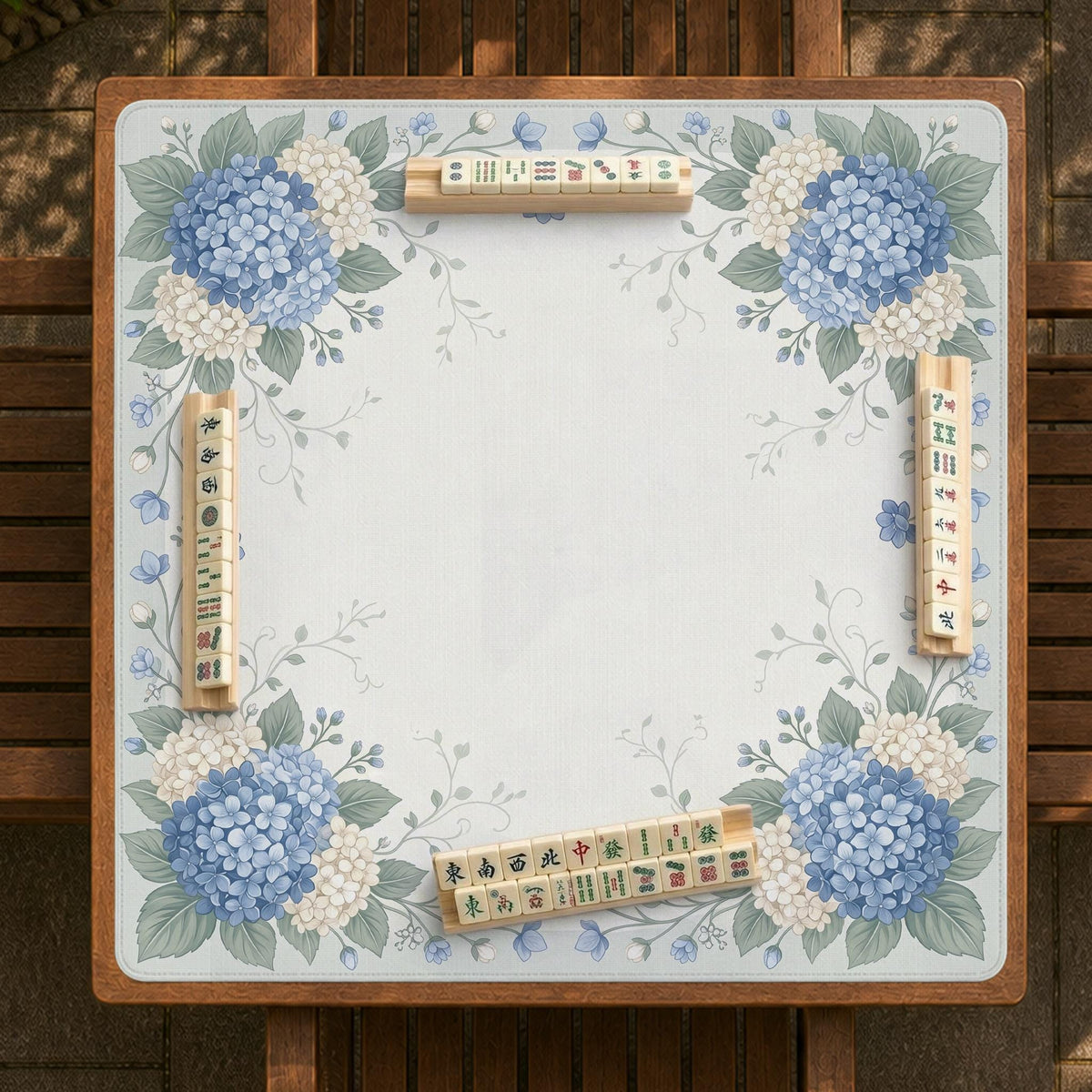 A square blue mahjong mat with hydrangea flowers, cream blossoms, sage leaves, and vines around a plain center play area. The edge is scalloped.