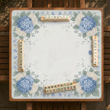 A square blue mahjong mat with hydrangea flowers, cream blossoms, sage leaves, and vines around a plain center play area. The edge is scalloped.