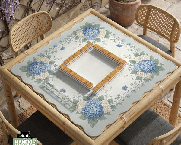 A square blue mahjong mat with hydrangea flowers, cream blossoms, sage leaves, and vines around a plain center play area. The edge is scalloped.