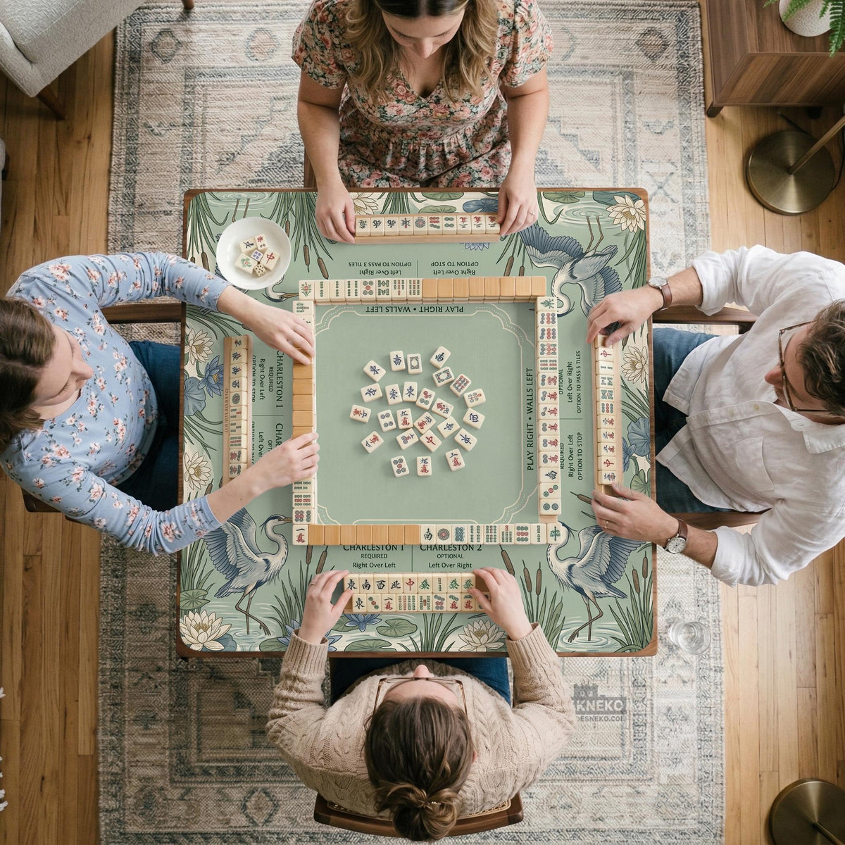 Square sage-green mahjong mat with blue-grey herons, water lilies, reeds, and printed play-direction and Charleston passing reminders around the playing area.