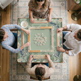 Square sage-green mahjong mat with blue-grey herons, water lilies, reeds, and printed play-direction and Charleston passing reminders around the playing area.