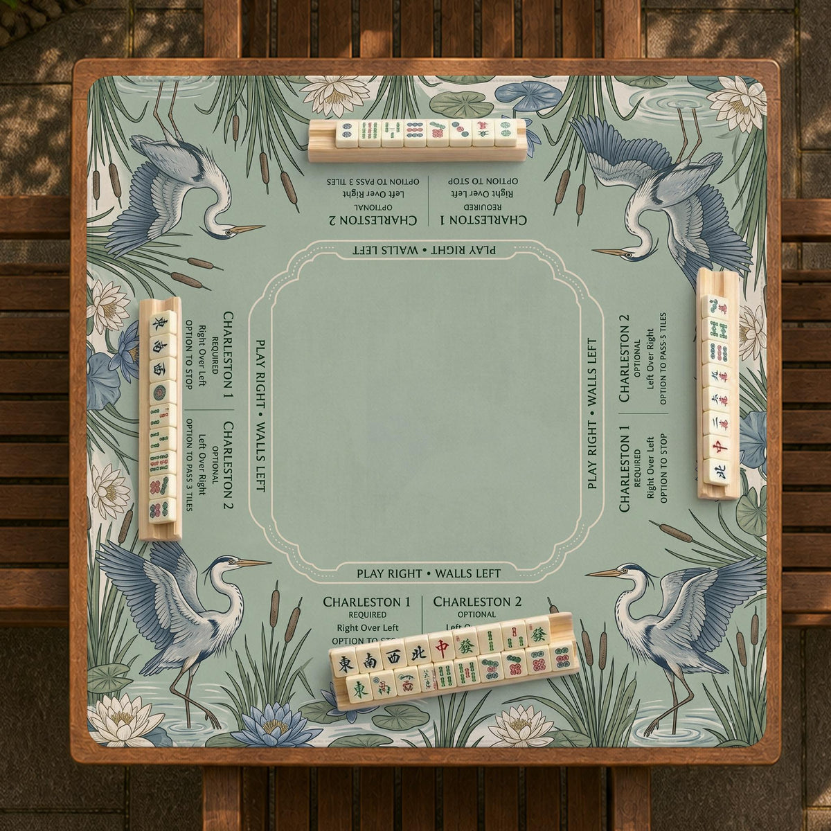 Square sage-green mahjong mat with blue-grey herons, water lilies, reeds, and printed play-direction and Charleston passing reminders around the playing area.