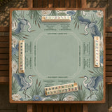 Square sage-green mahjong mat with blue-grey herons, water lilies, reeds, and printed play-direction and Charleston passing reminders around the playing area.