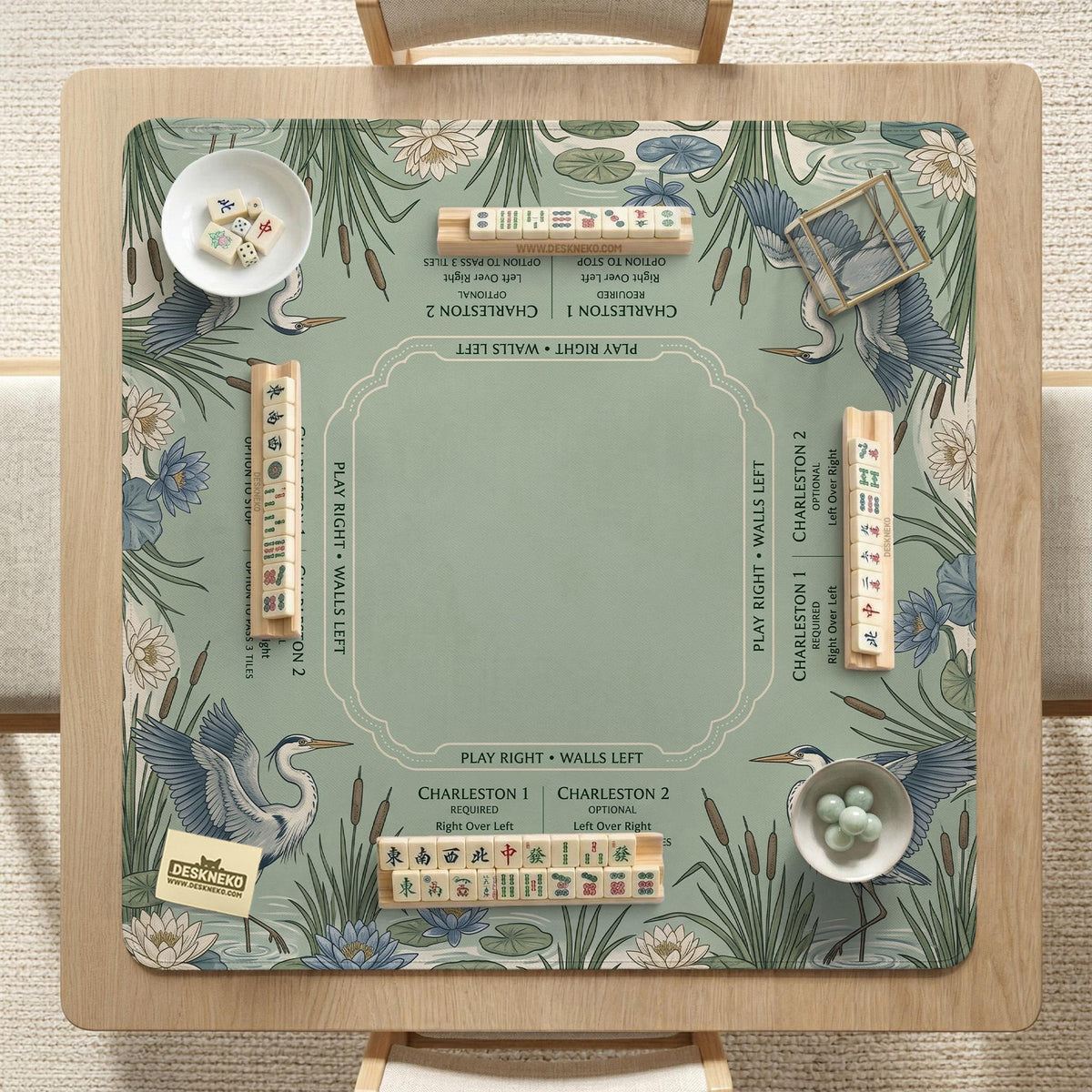 Square sage-green mahjong mat with blue-grey herons, water lilies, reeds, and printed play-direction and Charleston passing reminders around the playing area.