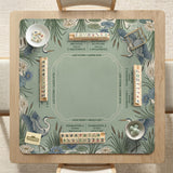 Square sage-green mahjong mat with blue-grey herons, water lilies, reeds, and printed play-direction and Charleston passing reminders around the playing area.
