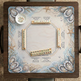 American mahjong mat with a cream center and a coastal border showing oyster shells, scallop shells, starfish, pearl strands, and pale blue sea plants.