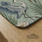 Square sage-green mahjong mat with blue-grey herons, water lilies, reeds, and printed play-direction and Charleston passing reminders around the playing area.