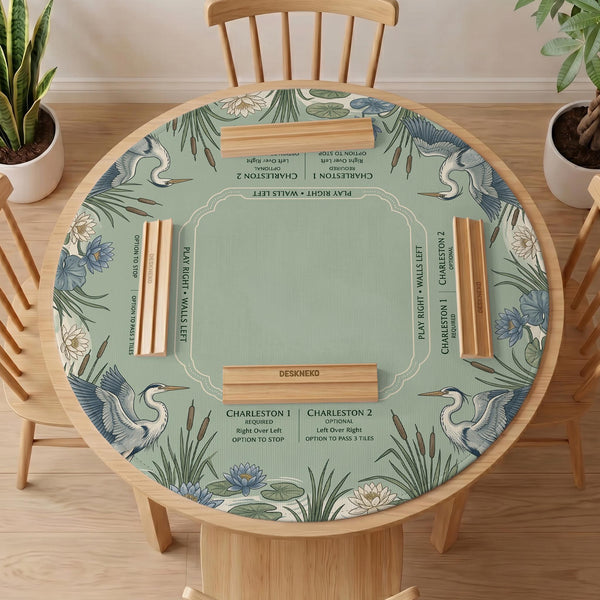 Square sage-green mahjong mat with blue-grey herons, water lilies, reeds, and printed play-direction and Charleston passing reminders around the playing area.