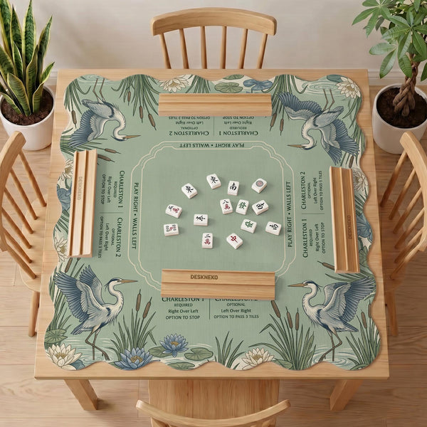 Square sage-green mahjong mat with blue-grey herons, water lilies, reeds, and printed play-direction and Charleston passing reminders around the playing area.