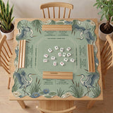 Square sage-green mahjong mat with blue-grey herons, water lilies, reeds, and printed play-direction and Charleston passing reminders around the playing area.