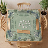 Square sage-green mahjong mat with blue-grey herons, water lilies, reeds, and printed play-direction and Charleston passing reminders around the playing area.