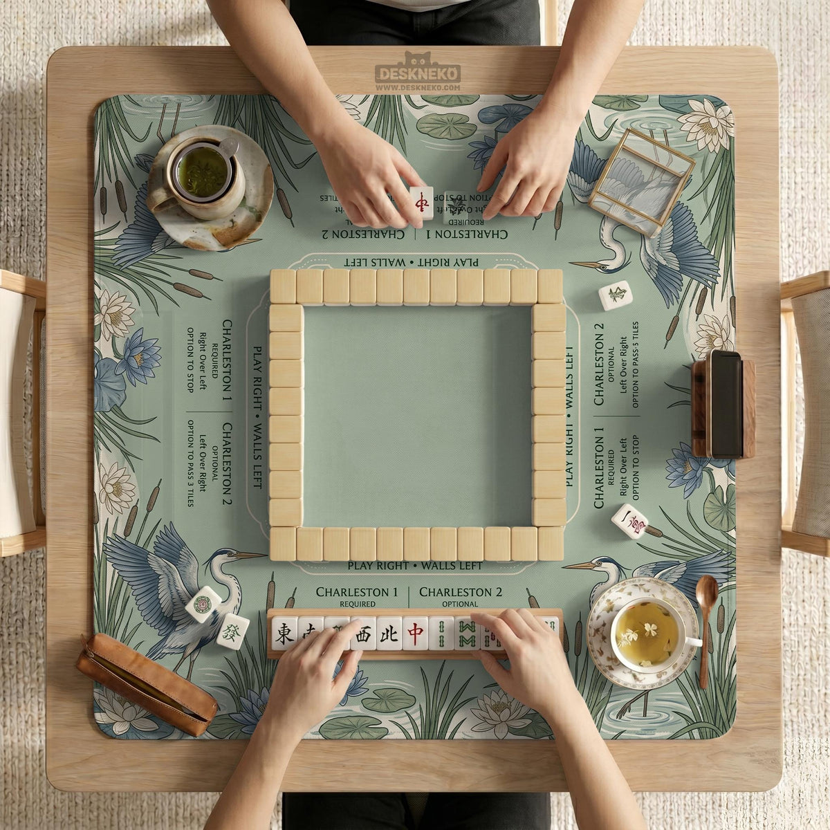 Square sage-green mahjong mat with blue-grey herons, water lilies, reeds, and printed play-direction and Charleston passing reminders around the playing area.