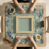 Square sage-green mahjong mat with blue-grey herons, water lilies, reeds, and printed play-direction and Charleston passing reminders around the playing area.