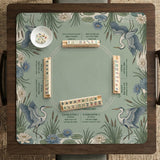 Square sage-green mahjong mat with blue-grey herons, water lilies, reeds, and printed play-direction and Charleston passing reminders around the playing area.