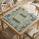 Square sage-green mahjong mat with blue-grey herons, water lilies, reeds, and printed play-direction and Charleston passing reminders around the playing area.